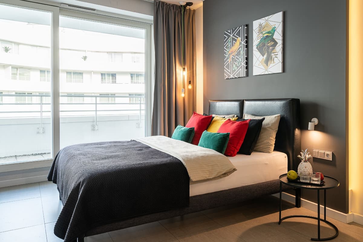 Une chambre à coucher moderne avec un grand lit, des coussins colorés, une table de chevet avec une lampe et des œuvres d’art accrochées au mur.