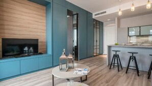 Ein modernes Wohnzimmer mit Fernseher, einem runden Tisch mit Gläsern und Äpfeln sowie einer Küchenzeile mit Hockern und einer Bar.