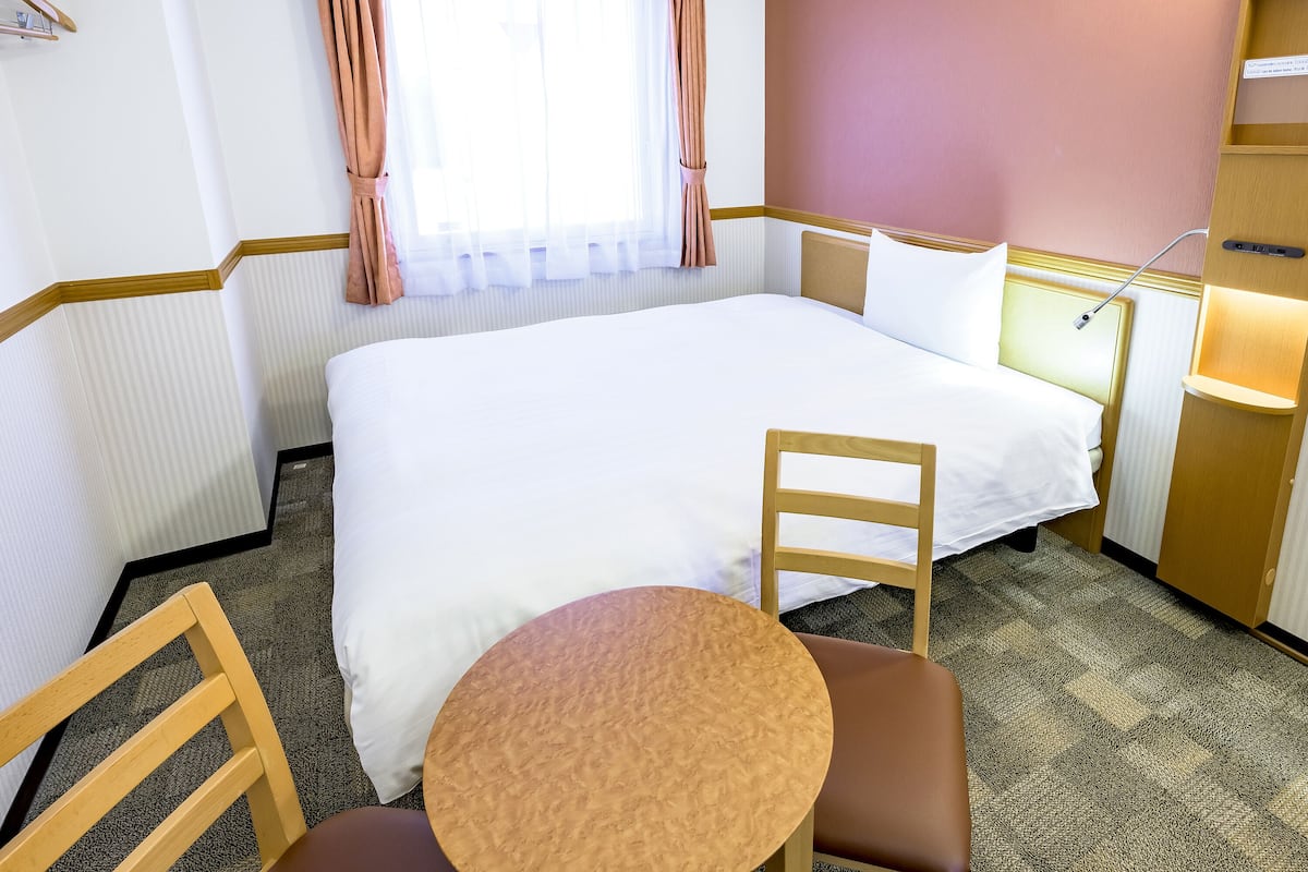Een hotelkamer met een groot bed, een houten stoel, een klein rond tafeltje en een nachtkastje met een lamp.