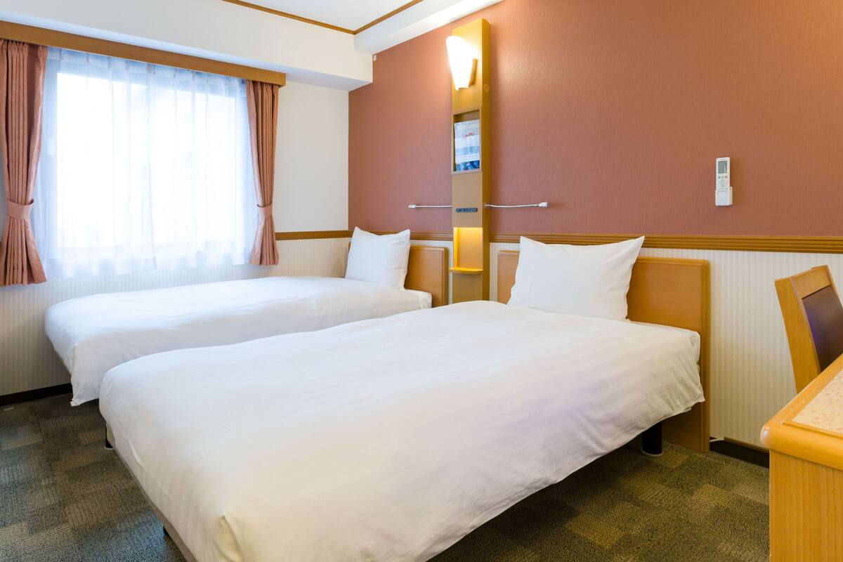 Een hotelkamer met twee bedden, een groot raam met gordijnen, een wandlamp en een klein bureau.