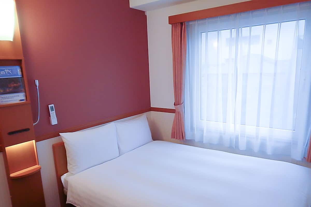 A hotel room with a bed, a bedside table, a nightstand, a window with curtains, and a wall-mounted thermostat.