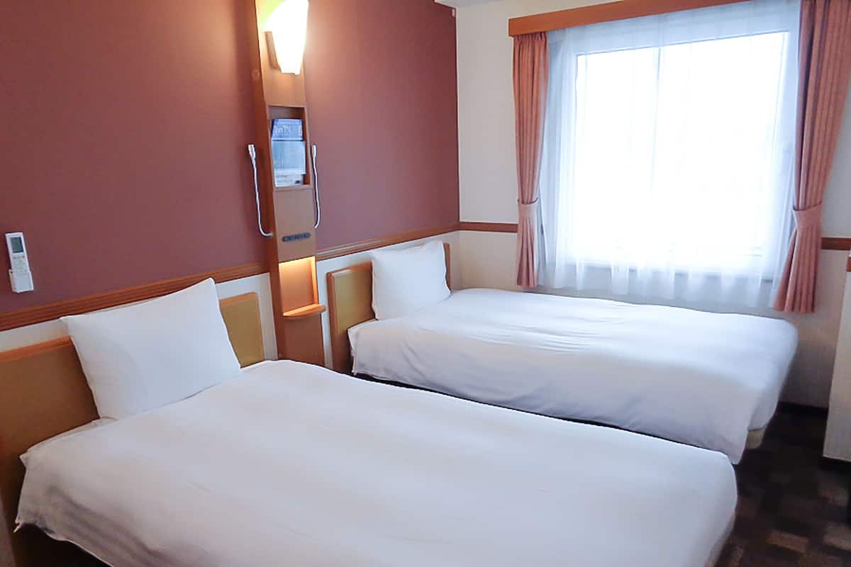 A hotel room with two beds, a bedside table with a lamp, a window with curtains, and a wall-mounted thermostat.