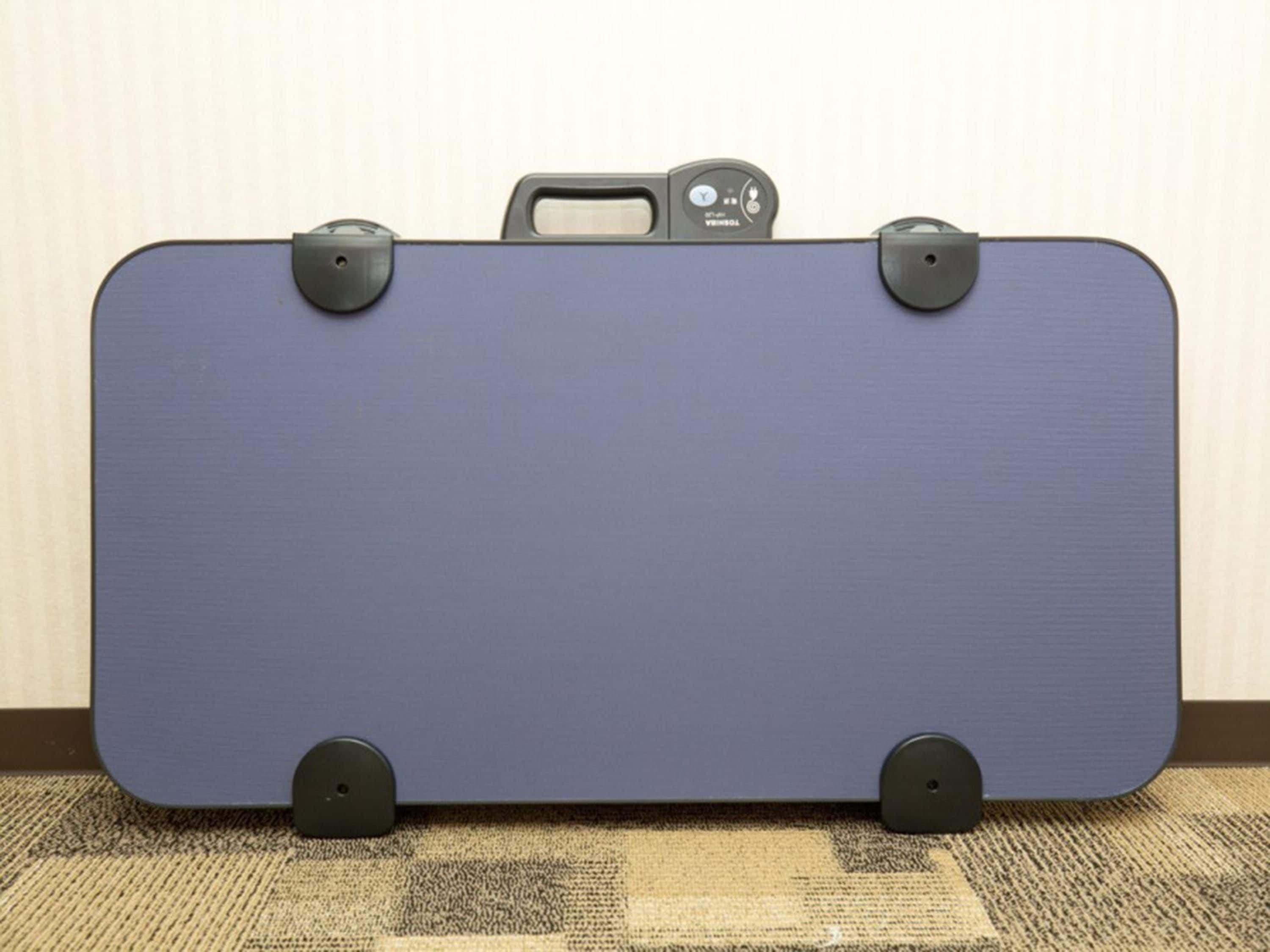 Une valise bleue avec des poignées et des roulettes noires, posée sur un sol recouvert d’une moquette, contre un mur.