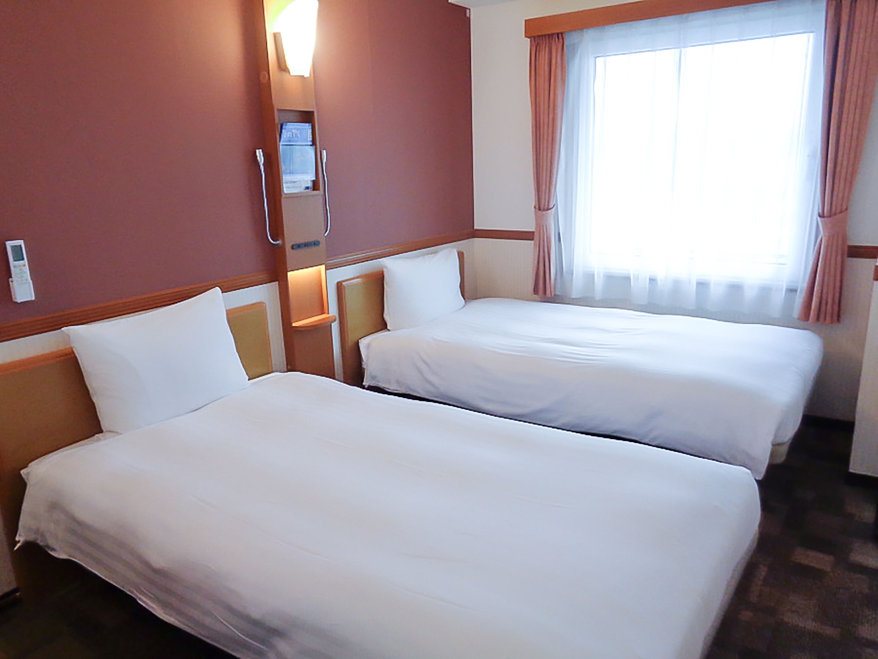 A hotel room with two beds, a bedside table with a lamp, a window with curtains, and a wall-mounted thermostat.
