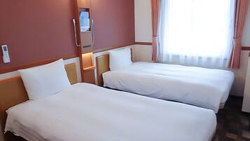 A hotel room with two beds, a bedside table with a lamp, a window with curtains, and a wall-mounted thermostat.