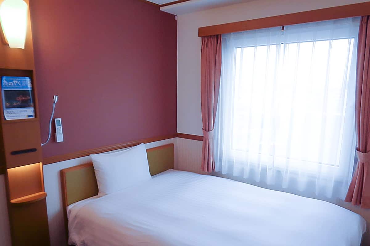 A hotel room with a single bed, a bedside table, a nightstand, a window with curtains, and a wall-mounted phone.