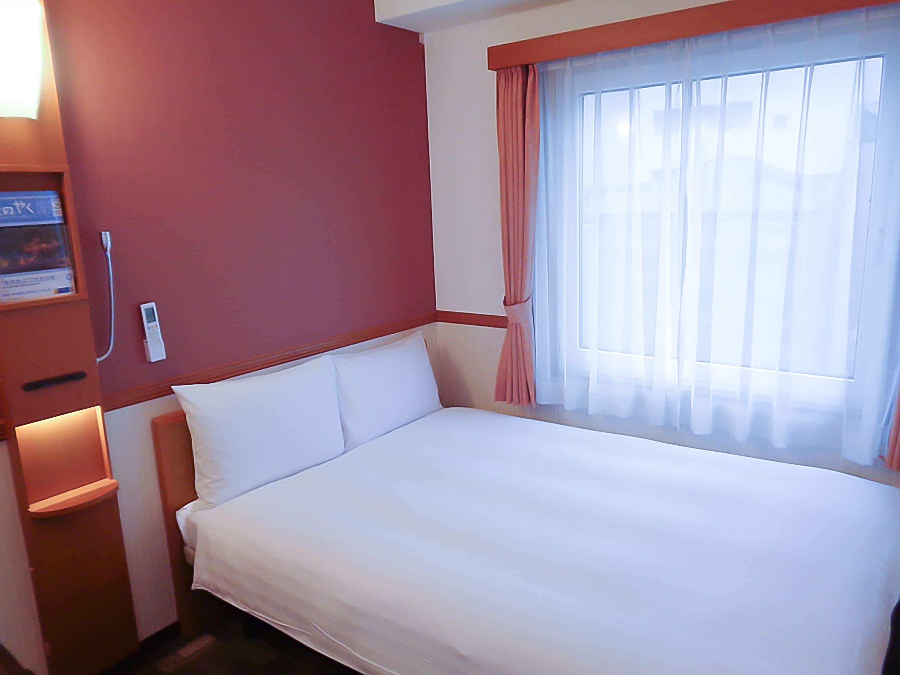 A hotel room with a bed, a bedside table, a nightstand, a window with curtains, and a wall-mounted thermostat.