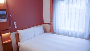 A hotel room with a bed, a bedside table, a nightstand, a window with curtains, and a wall-mounted thermostat.