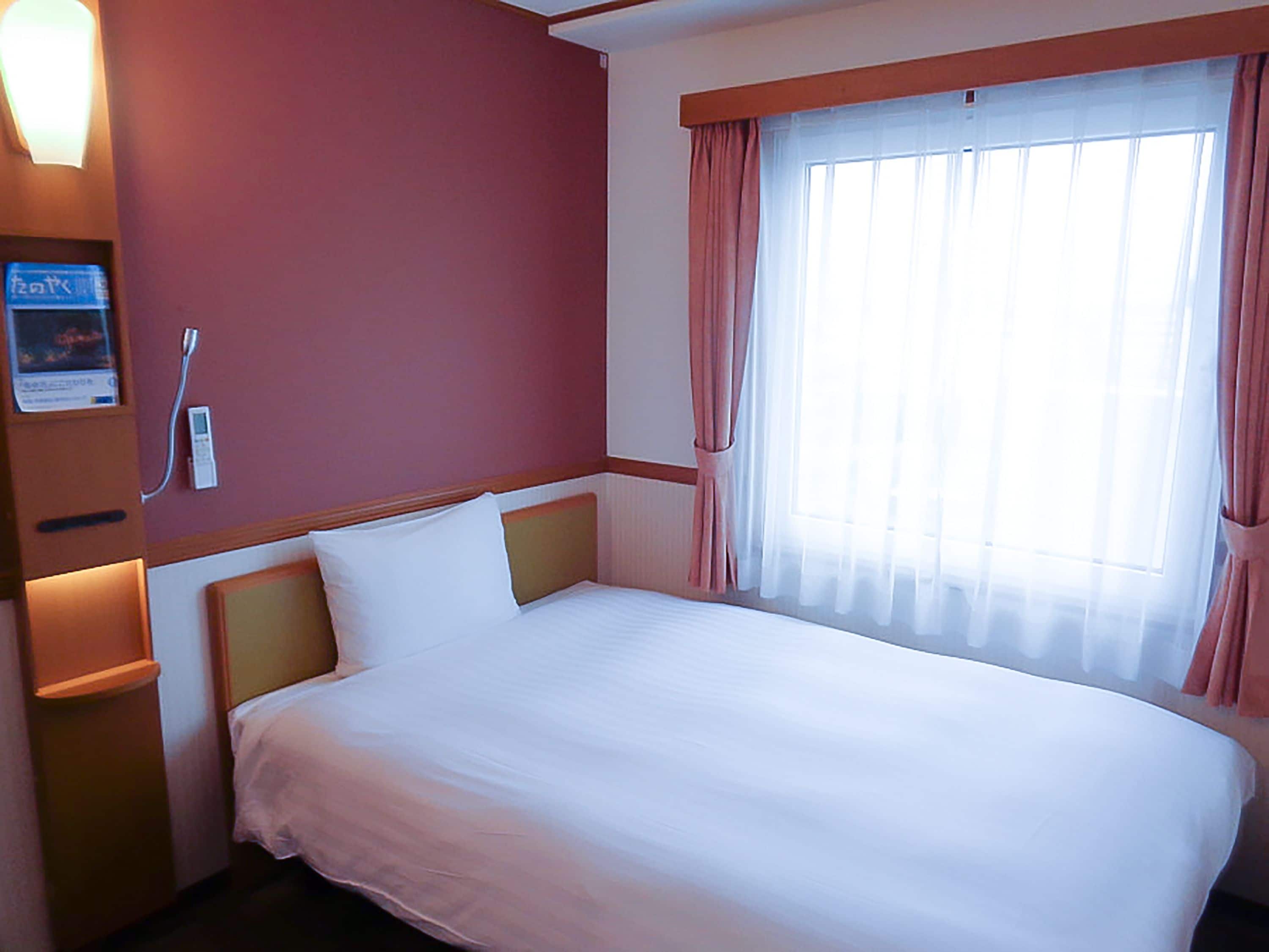A hotel room with a single bed, a bedside table, a nightstand, a window with curtains, and a wall-mounted phone.