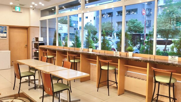 Un espace de détente intérieur moderne, avec des tables et des chaises, de grandes fenêtres offrant une vue sur la rue et les bâtiments environnants.
