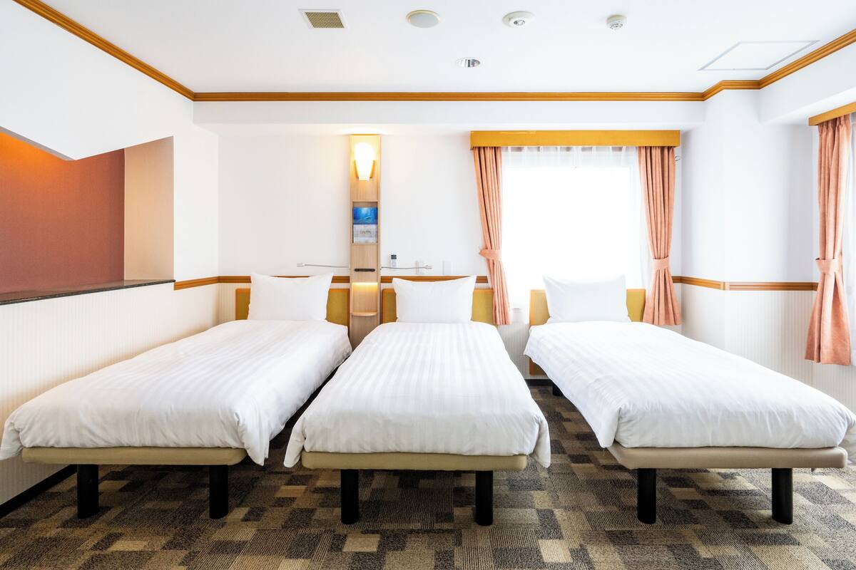 A hotel room with two single beds, a wooden headboard, a wall-mounted TV, and a window with curtains.