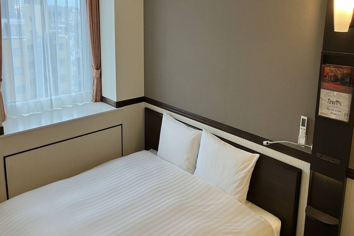 Ein Hotelzimmer mit einem Bett, einem Fenster mit Vorhängen, einem an der Wand befestigten Thermostat und einem kleinen Bilderrahmen.
