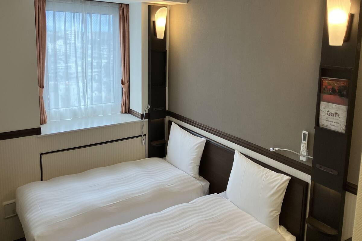 Ein Hotelzimmer mit zwei Betten, einem Fenster mit Vorhängen, einer Wandleuchte und einem digitalen Gerät an der Wand.