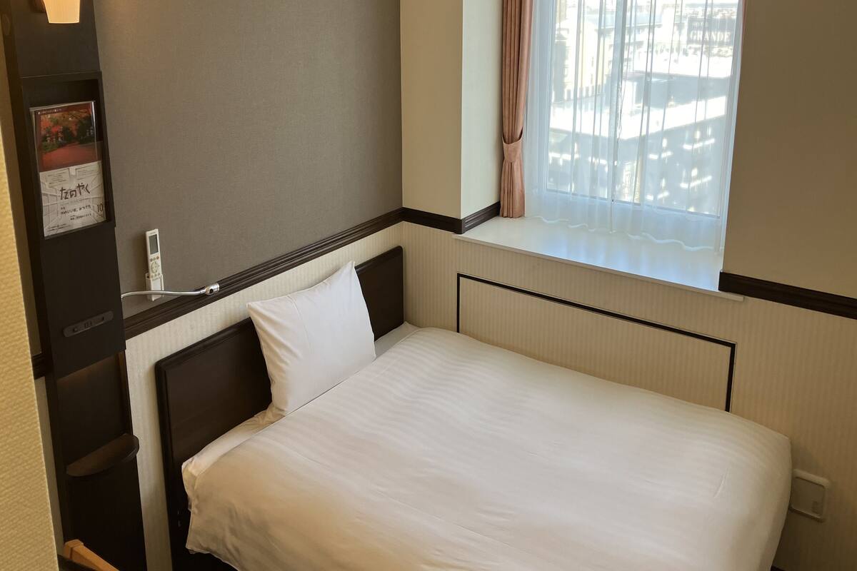 Ein Einzelbett mit weißer Matratze und Kissen, ein Nachttisch mit Lampe, ein Fenster mit Blick auf Gebäude und ein an der Wand befestigtes Thermostat.
