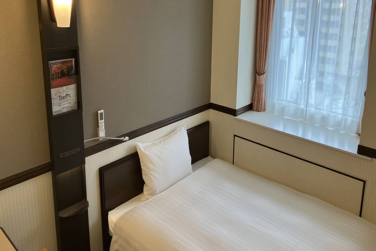 Ein Hotelzimmer mit einem Einzelbett, einem Nachttisch, Vorhängen an einem Fenster und einem an der Wand befestigten Thermostat.