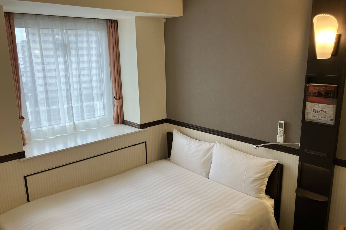 Ein Hotelzimmer mit einem großen Bett, einem Fenster mit Vorhängen, einer Wandleuchte und einem digitalen Gerät an der Wand.