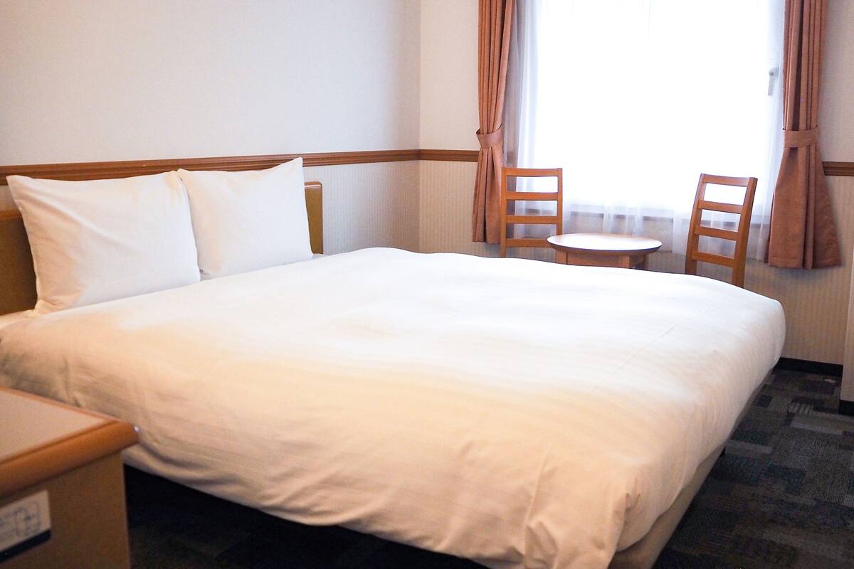 A hotel room with a large bed, two wooden chairs, a small round table, and a window with curtains.