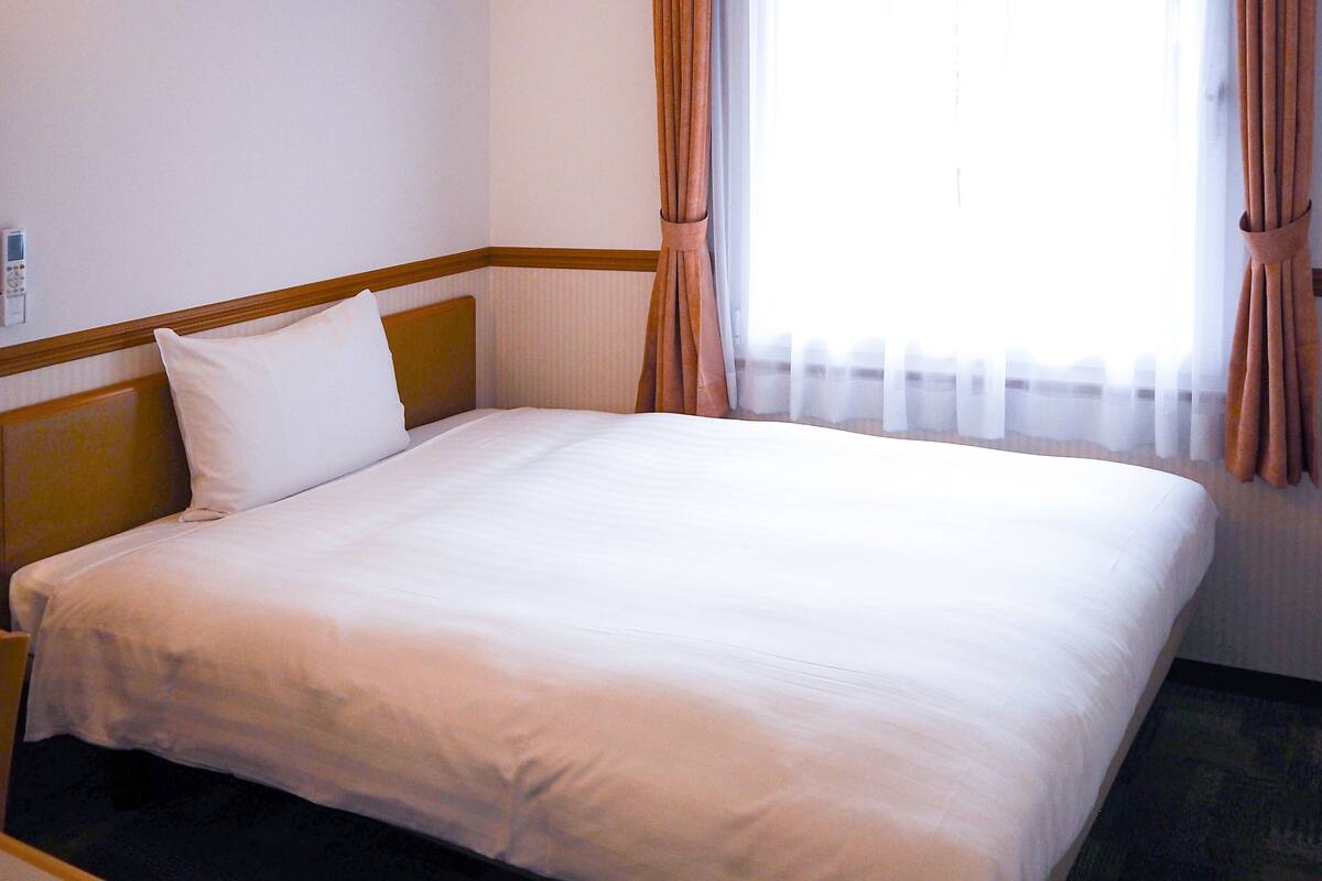 A neatly made bed in a hotel room with a wooden headboard, a bedside table, and a window with curtains.
