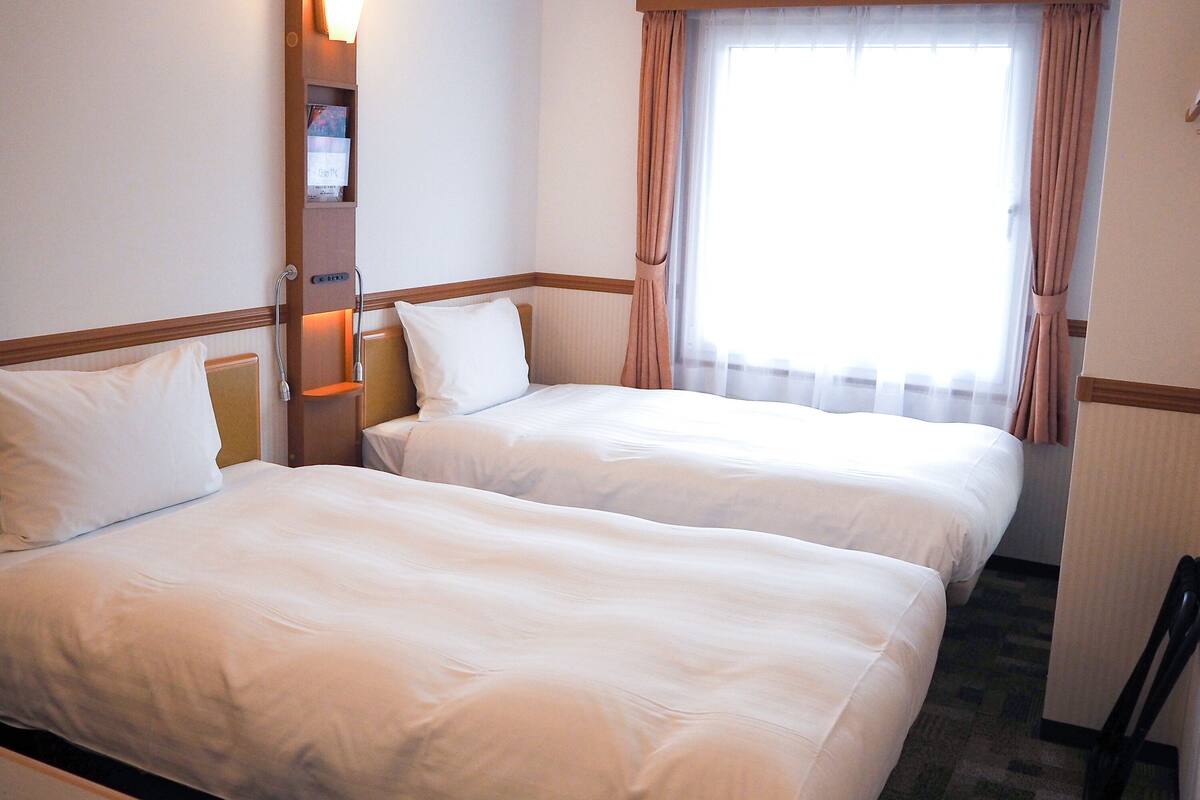 A hotel room with two beds, a nightstand, a lamp, and a window with curtains.