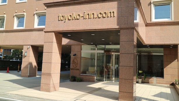 A hotel entrance with a clear glass door, a sign reading "toyoko-inn.com", and a tiled pavement outside.