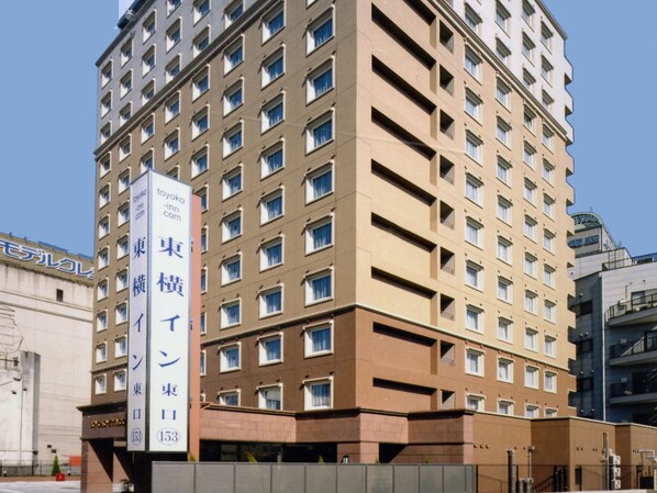 Un hôtel à plusieurs étages, doté d’une façade beige et de nombreuses fenêtres. Une enseigne verticale sur le bâtiment indique le nom de l’hôtel et son adresse sur le site web.