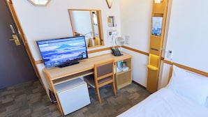 A hotel room with a bed, a desk with a computer, a small refrigerator, a bedside table with a lamp, and a mirror.