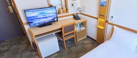 Une chambre d’hôtel comprenant un lit, un bureau avec un ordinateur, un petit réfrigérateur, une table de chevet avec une lampe et un miroir.