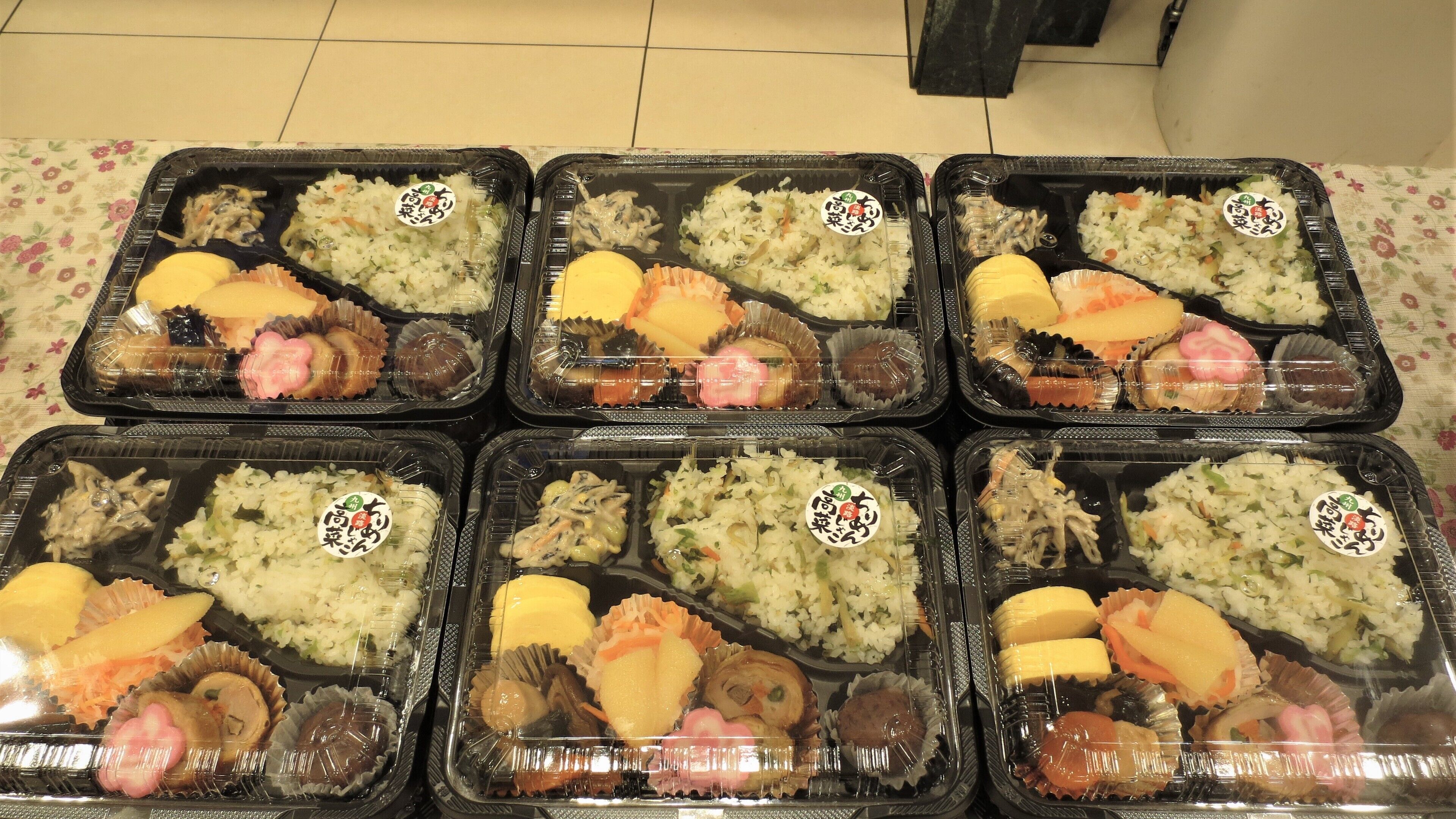 Six boîtes contenant des repas composés de riz, de légumes et de différentes sortes de viande, posées sur un sol carrelé.