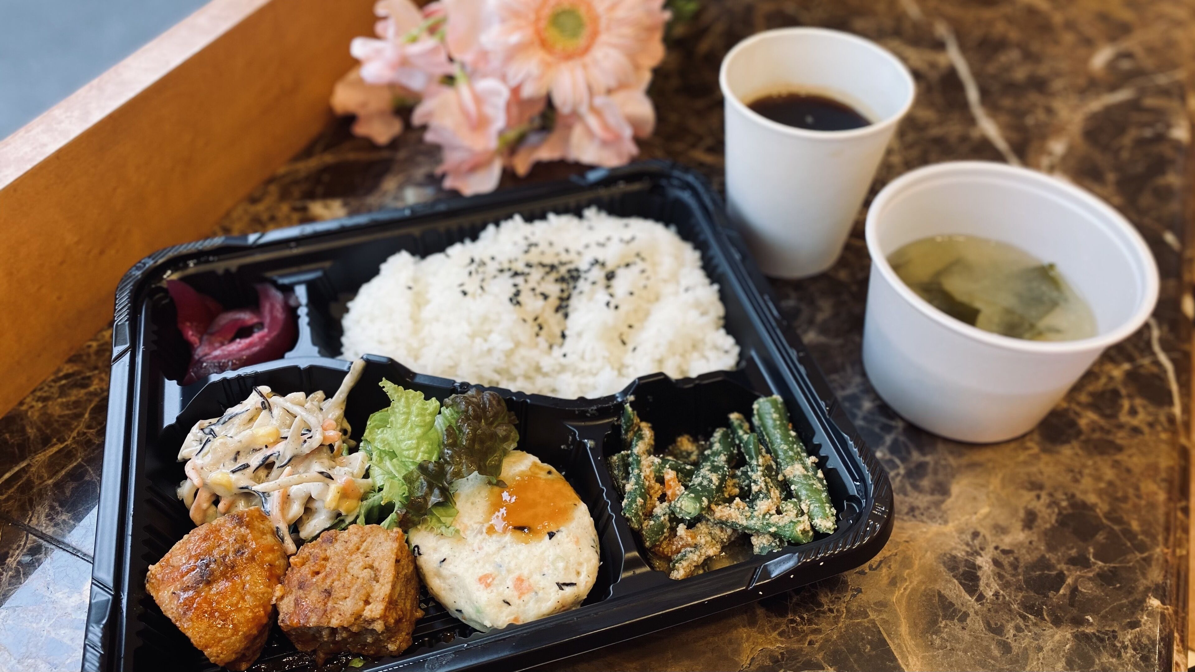 A meal in a black tray with rice, vegetables, and meat, accompanied by two cups of beverages and a flower.