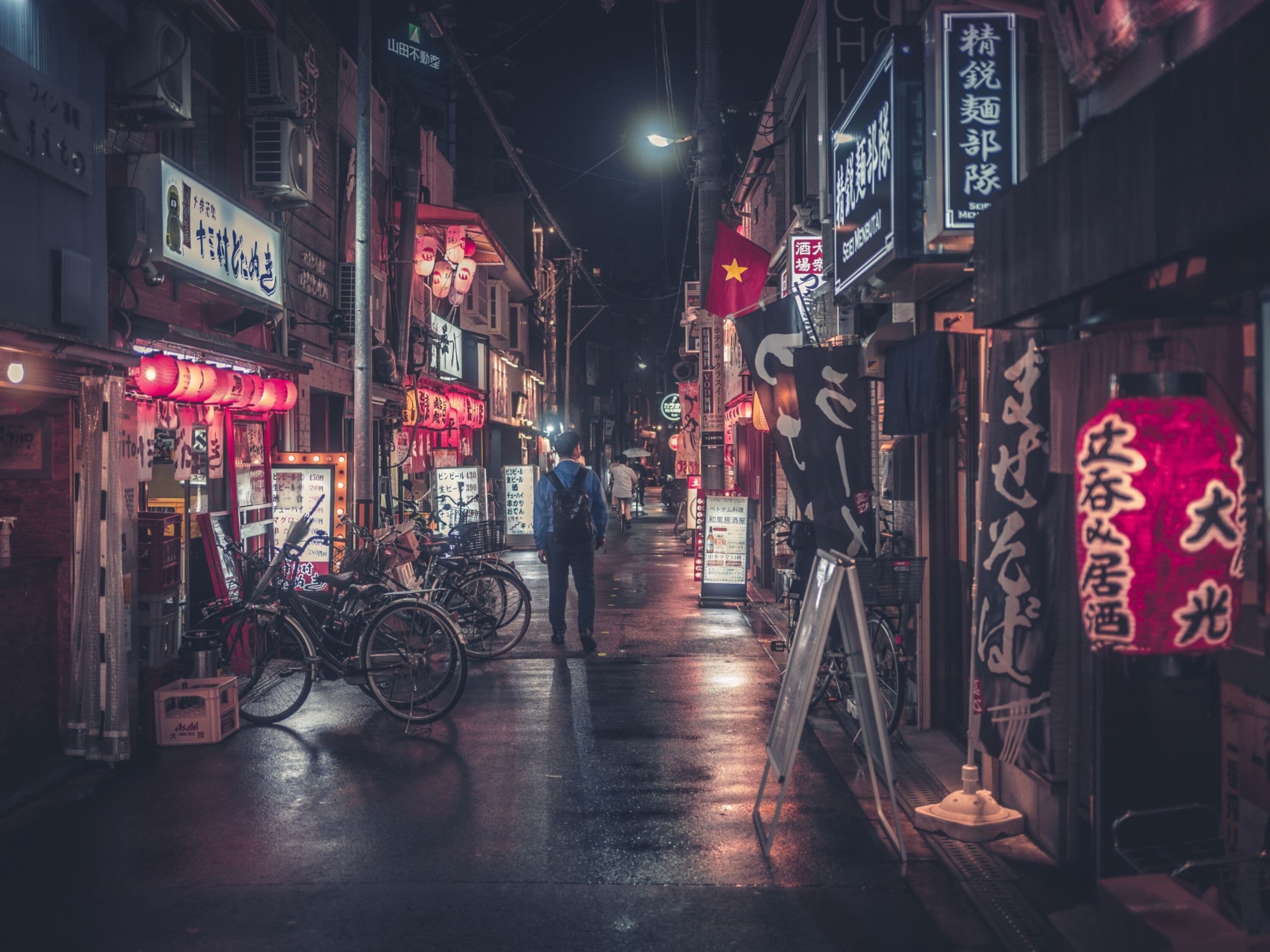 A narrow street at night with illuminated signs and storefronts, bicycles parked along the side, and a person walking down the street.