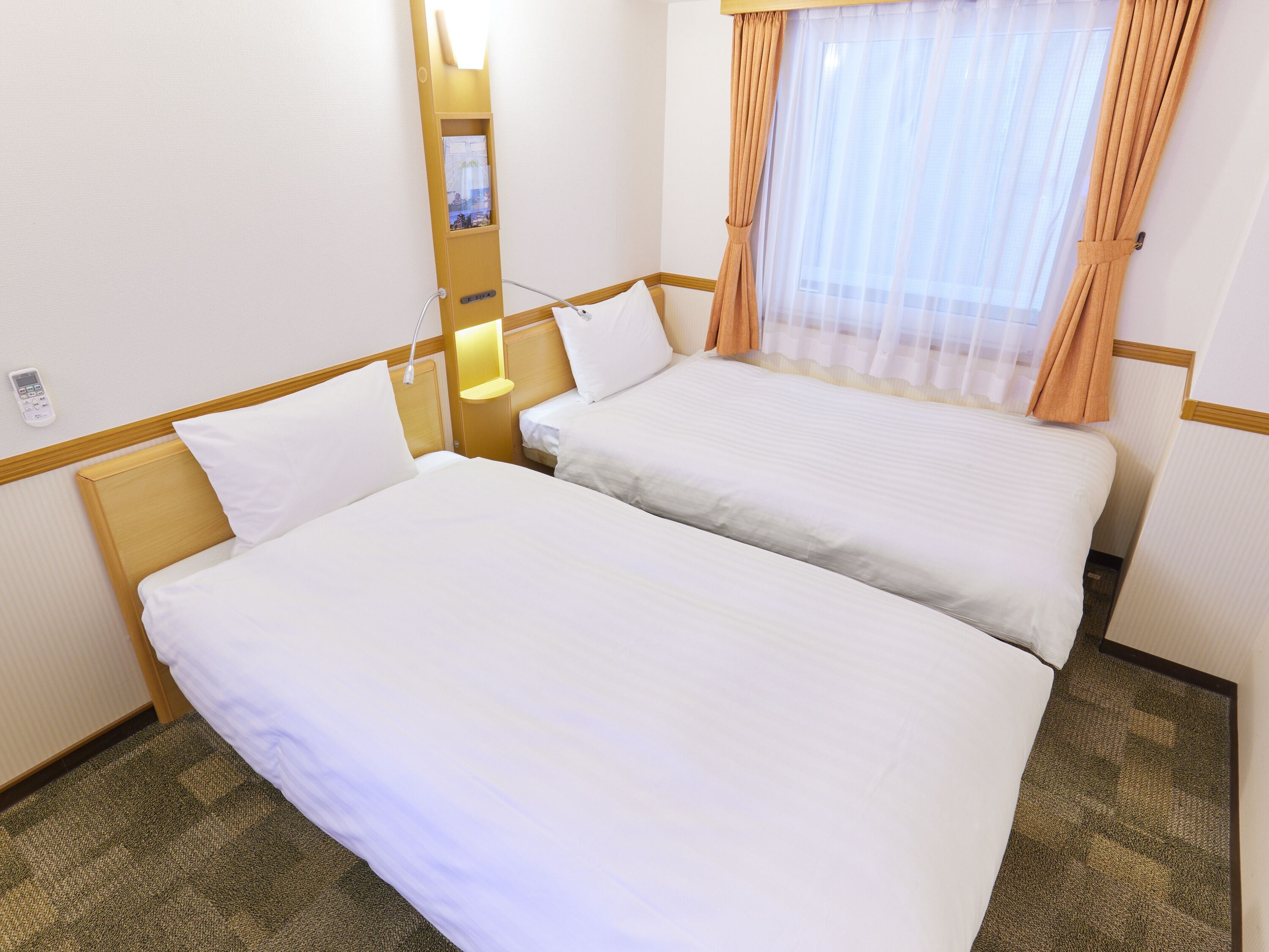 Hotelkamer met twee bedden, een nachtkastje met lamp, een raam met gordijnen en een telefoon aan de muur.