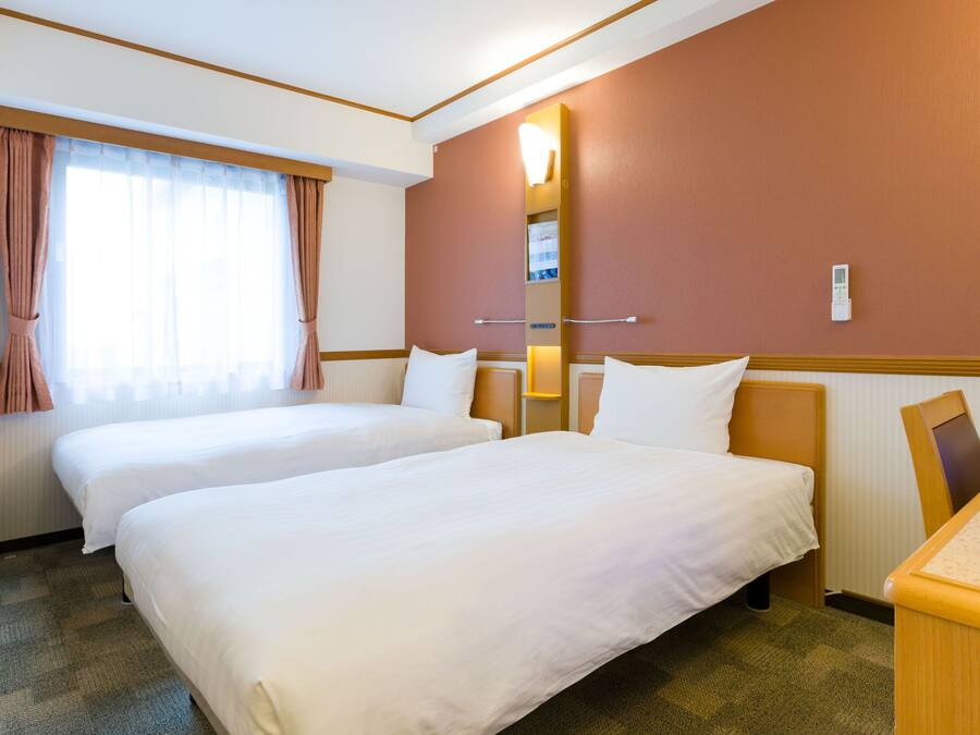 A hotel room with two beds, a large window with curtains, a wall-mounted light, and a small desk.