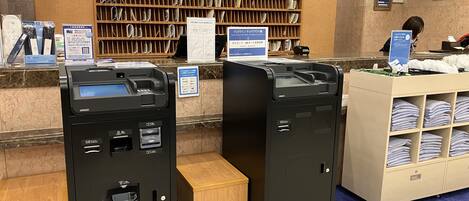 A hotel reception area with a wooden reception desk, a clock, and a shelf with folded towels. There are two ATMs and a small wooden bench.