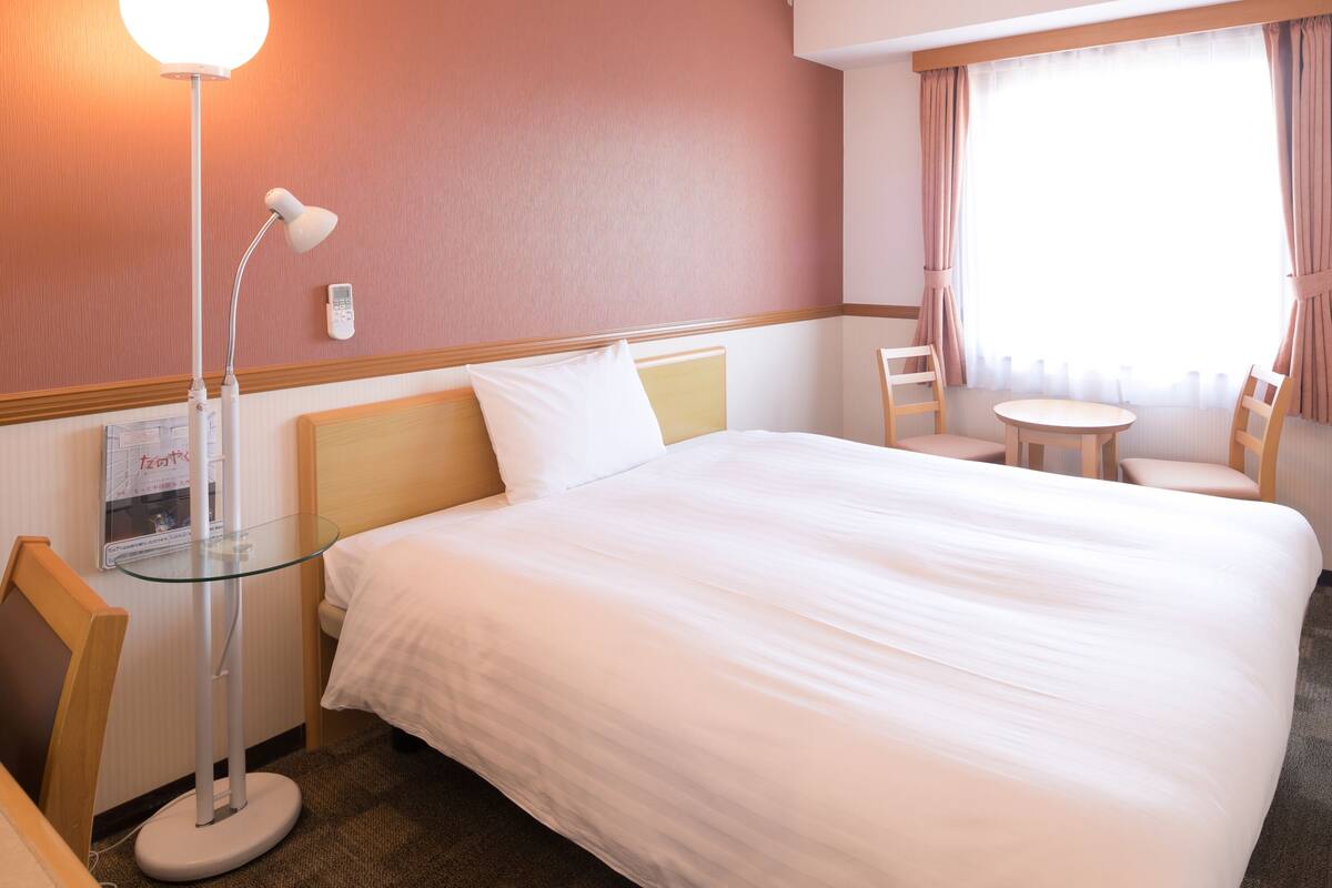 A hotel room with a large bed, a bedside table, a lamp, a chair, a small table, and a window with curtains.