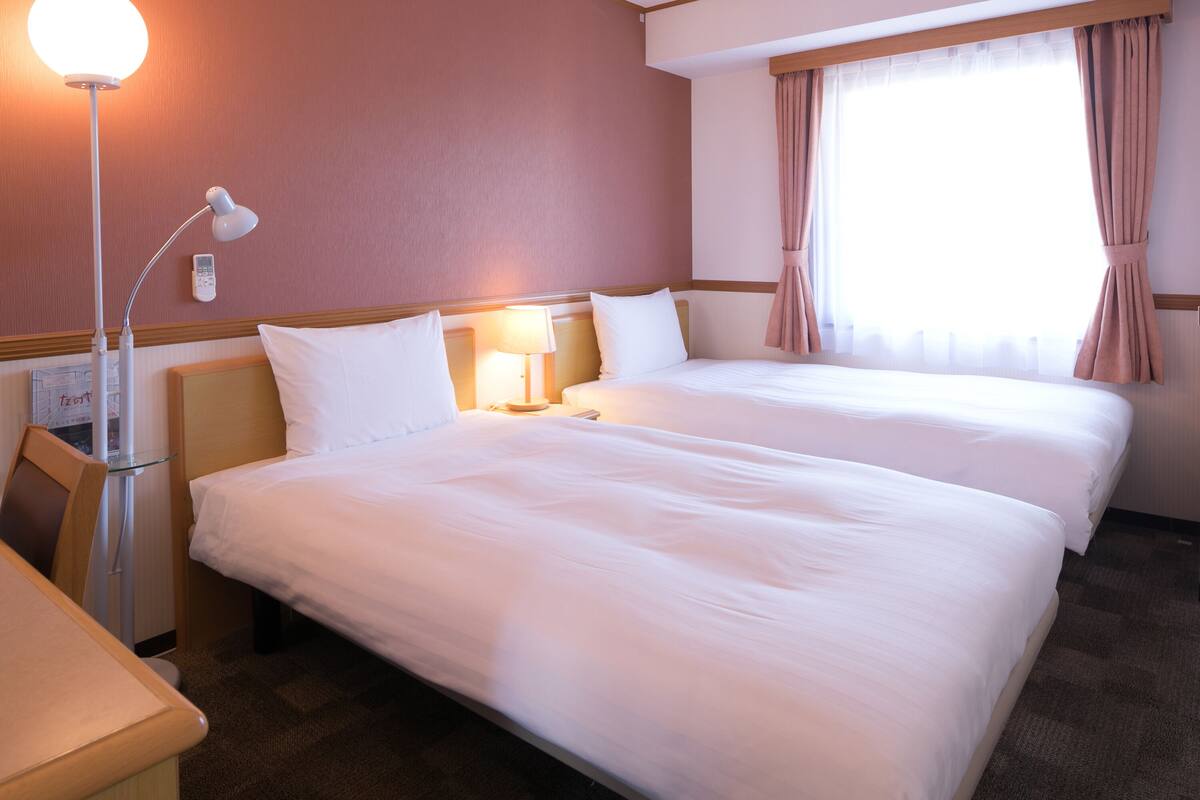 A hotel room with two beds, a desk, a lamp, and a window with curtains.
