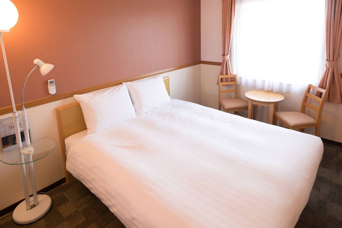 A hotel room with a large bed, a bedside lamp, a small table with a chair, and a window with curtains.