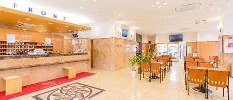 Zona de recepción de un hotel, con mostrador de recepción, área de asientos, máquinas expendedoras y una pantalla grande.