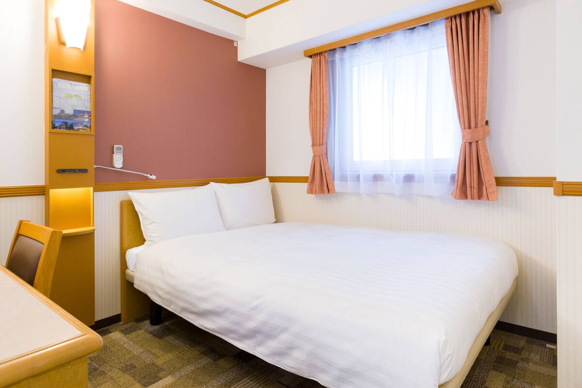 A hotel room with a large bed, a desk, a chair, a bedside table, a wall-mounted phone, and a window with curtains.
