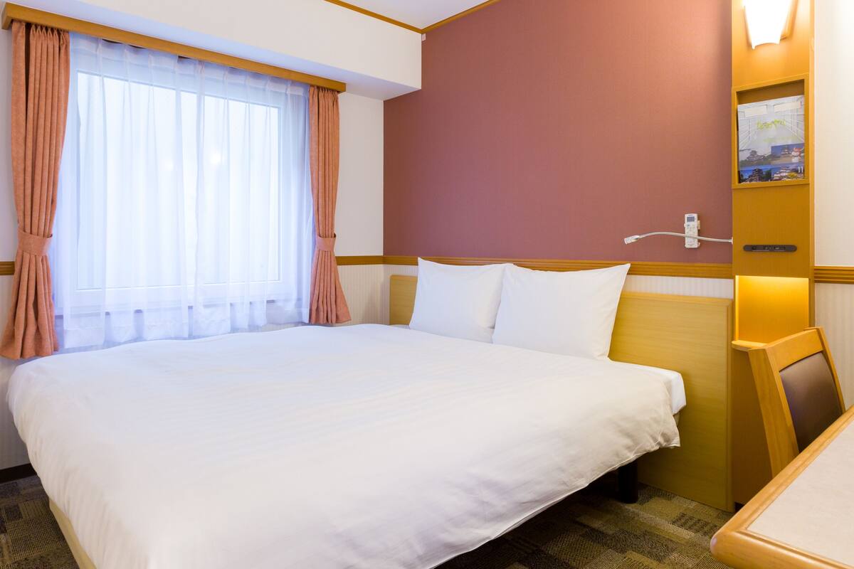 A hotel room with a large bed, a wooden headboard, a window with sheer curtains, and a wall-mounted light fixture.