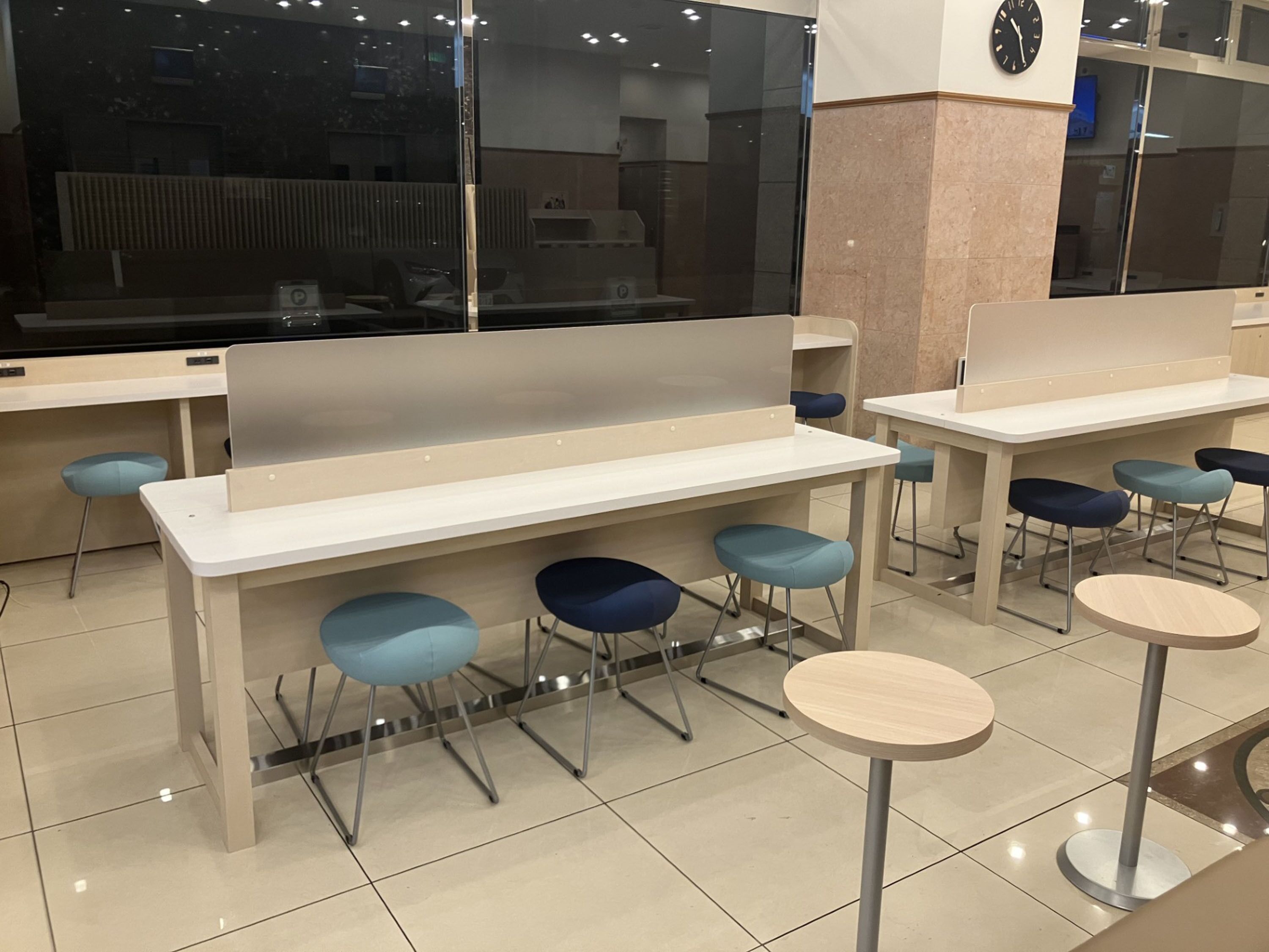 Una cafetería moderna con mesas altas y sillas azules, un reloj en la pared y un amplio ventanal.