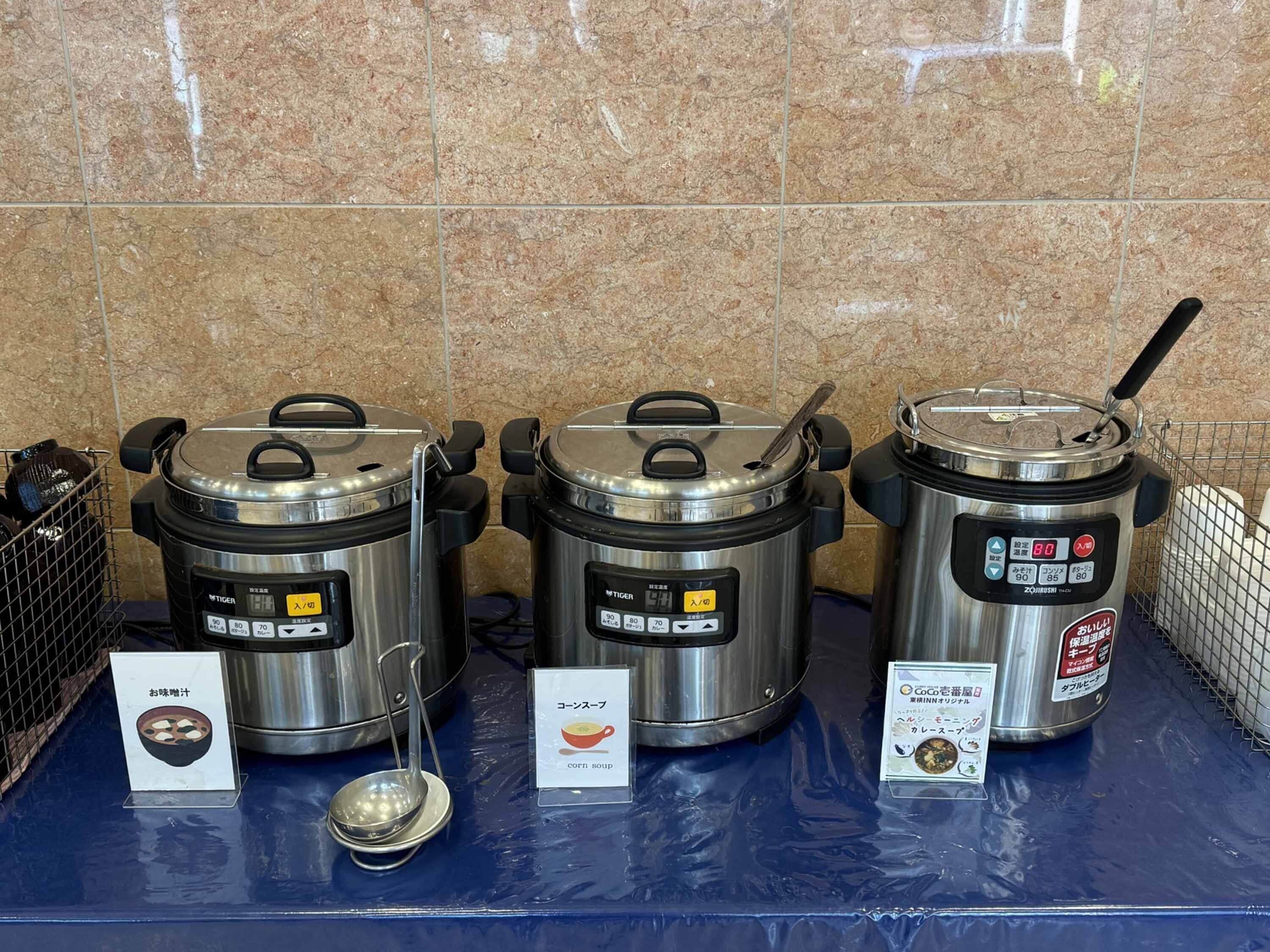 Drie roestvrijstalen slowcookers met deksels, geplaatst op een blauw oppervlak met labels die aangeven dat ze geschikt zijn voor het maken van rijst- en maissoep.