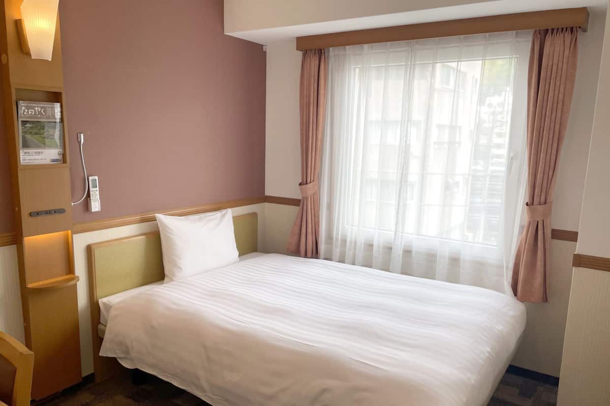 Una habitación de hotel con cama, mesita de noche, lámpara y ventana con cortinas.