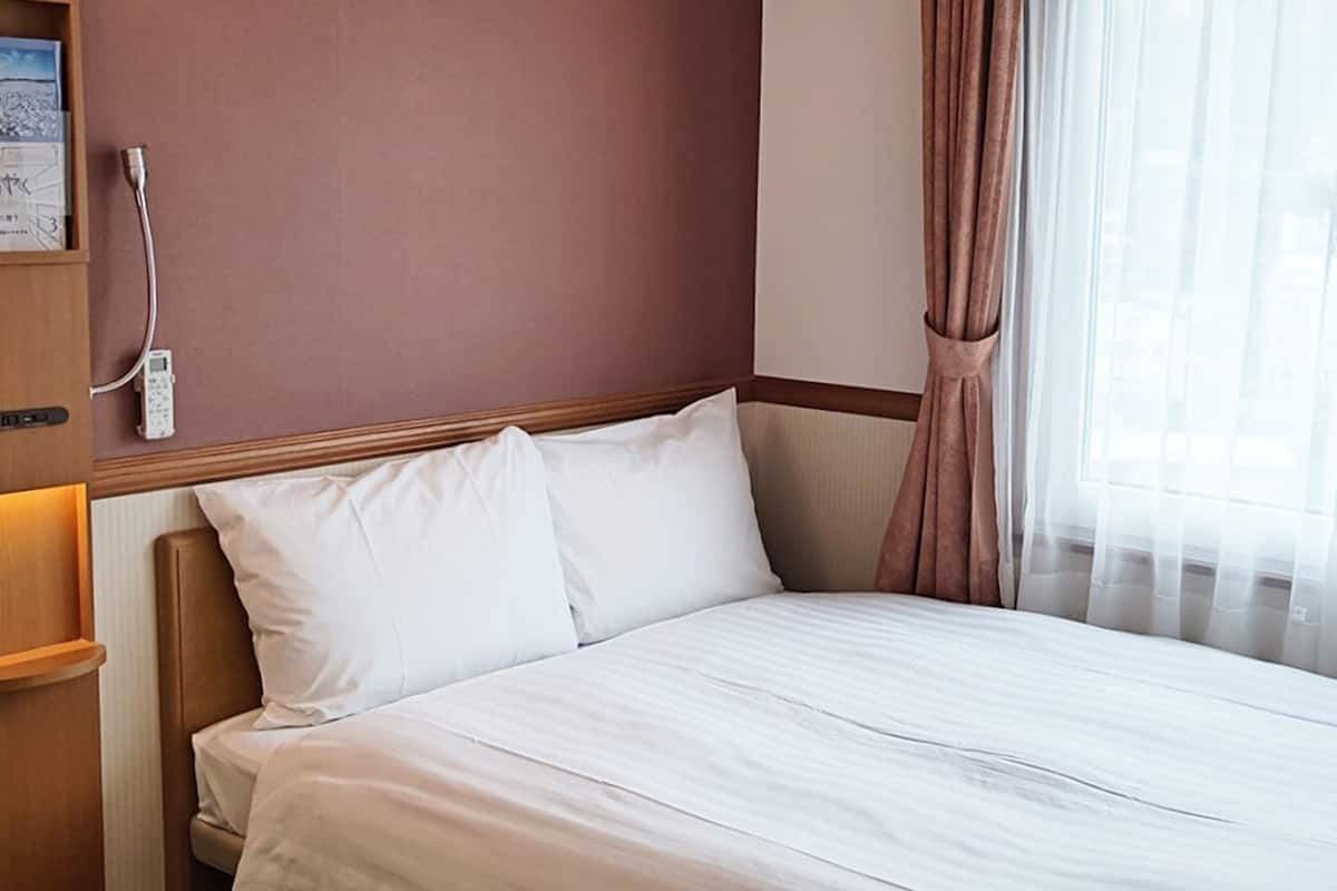 Una habitación de hotel con cama, mesita de noche, lámpara y ventana con cortinas.