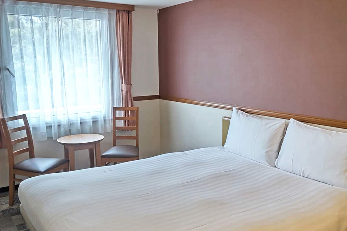Habitación de hotel con una cama grande, una mesita redonda pequeña, dos sillas y una ventana con cortinas de gasa.