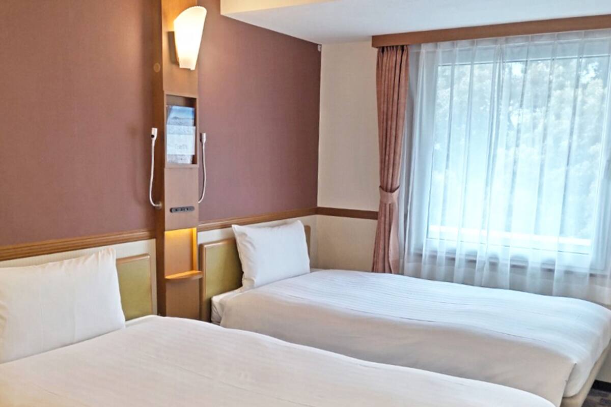 Una habitación de hotel con dos camas, una ventana con cortinas traslúcidas y una lámpara fijada a la pared.