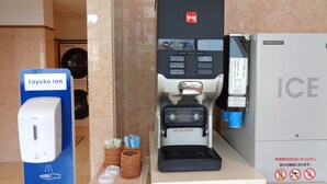 Une machine à café dotée d’un panneau de commande et d’un distributeur de tasses, un panneau indiquant « Toyoko Inn », et un distributeur de gel hydroalcoolique.