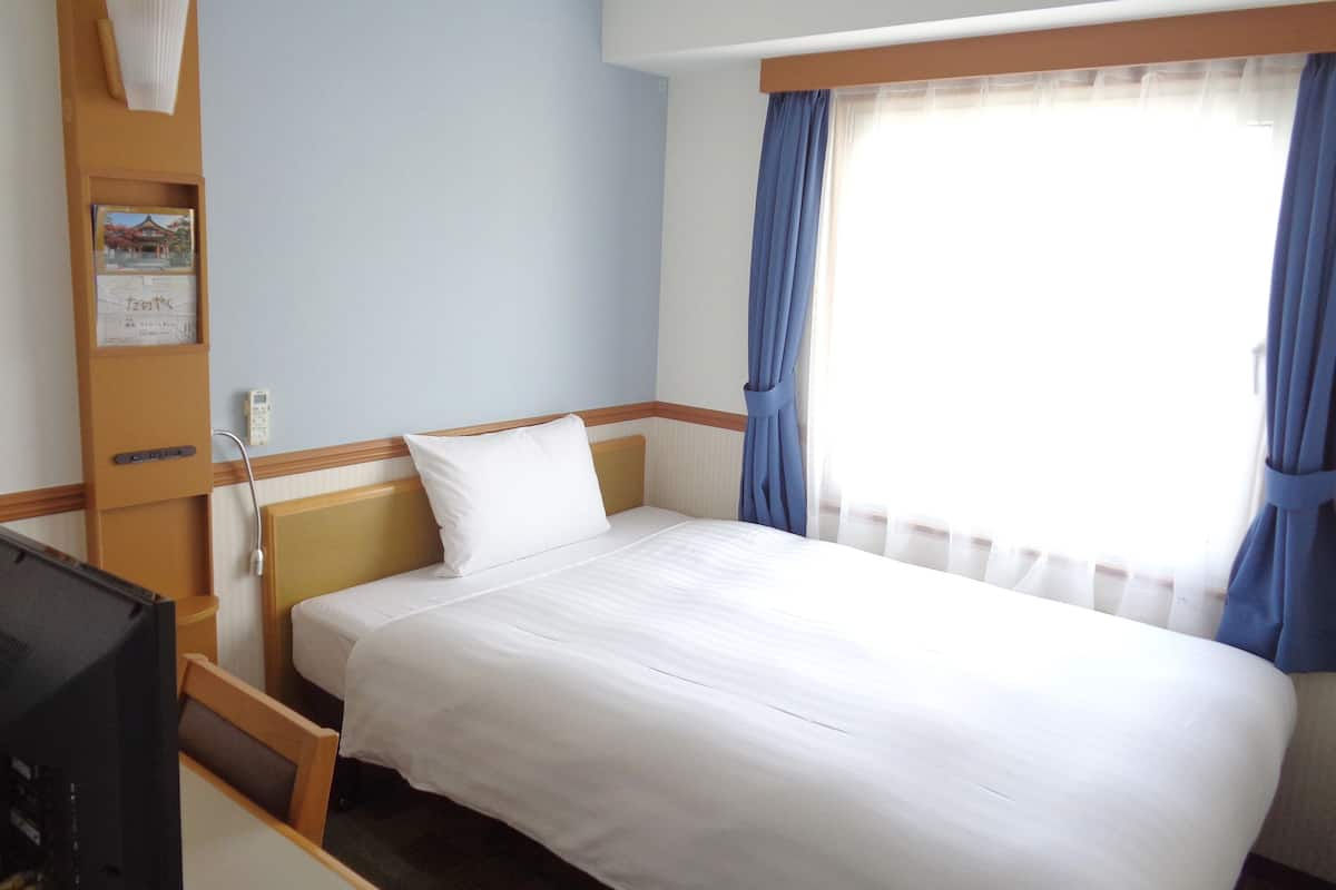 Hotelkamer met een bed, bureau, stoel, televisie, raam met gordijnen en een wandplank met een boek en decoratie.