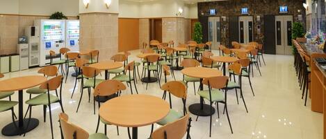 Una amplia cafetería con varias mesas y sillas redondas, máquinas expendedoras y un área de recepción con pared de mármol y pantallas de televisión.