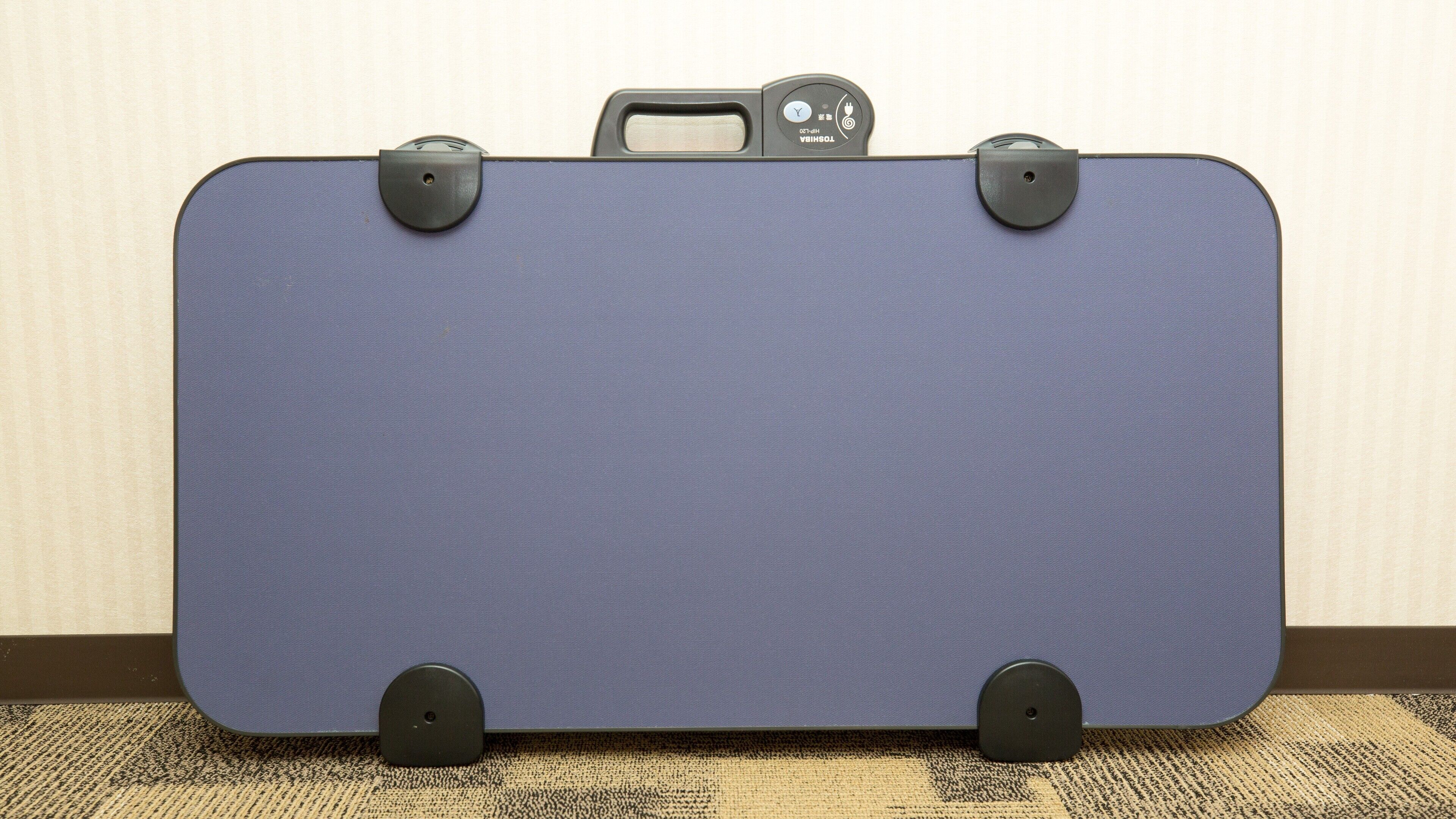 A blue suitcase with a handle and wheels, placed on a carpeted floor against a wall.