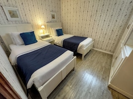 A room with two beds, each with a white headboard and blue bedding. There is a nightstand with a lamp between the beds. The walls have a floral pattern, and there is a framed picture above the nightstand.